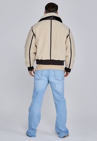 Beige fleece jacket with a black collar and trim, featuring a quilted back and ribbed cuffs, paired with light blue jeans.