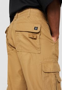 Homme portant un short cargo beige avec plusieurs poches à rabats et une chemise noire, main reposant dans la poche latérale.