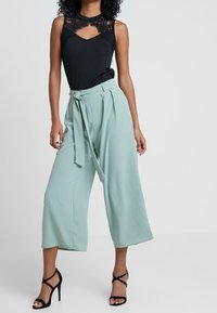 Haut noir sans manches avec des accents en dentelle, associé à un pantalon taille haute à jambes larges de couleur vert clair et des talons noirs à lanières.