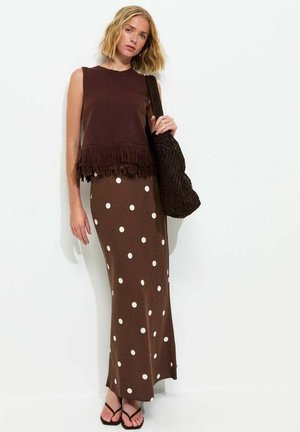 Vrouw die een mouwloos bruin topje met franjes draagt, een lange bruine rok met witte stippen, zwarte sandalen en een grote geweven bruine tas vasthoudt.