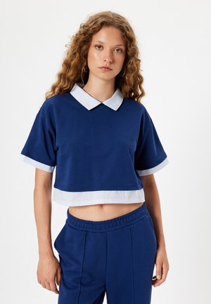 Felpa blu tagliata con colletto e polsini bianchi a strisce, presenta una texture liscia. Abbinata a pantaloni blu con vita elastica.