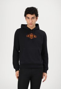 Svart hettegenser med oransje brodert logo på brystet, med frontlomme, snor i hetten og ribbestrikkede mansjetter. Glatt tekstur.