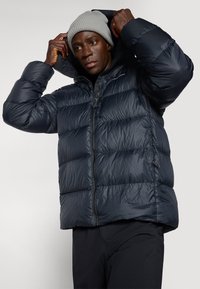 Veste matelassée bleu marine avec un design en piqué, fermeture éclair à l'avant et capuche. Le matériau extérieur est brillant, suggérant une isolation pour les temps froids.
