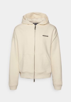 Bluza z kapturem na zamek w kremowym kolorze z przednimi kieszeniami oraz logo "WRSTBHVR" na lewej piersi, z kapturem ze sznurkiem i ściągaczami na rękawach.
