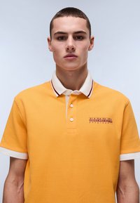 Orange polo-shirt i struktureret stof, hvid krave med røde detaljer, to-knap lukning og broderet logo på brystet.