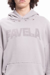 Cilvēks ar cirtainiem matiem, ģērbies gaiši pelēkā kapucē ar nolietotiem detaļām un uz krūšu ir izdrukāts vārds "FAVELA".