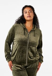 Felpa con zip color verde oliva in velour, dotata di tasca frontale, cappuccio con coulisse e polsini a costine, con una finitura morbida e texture.