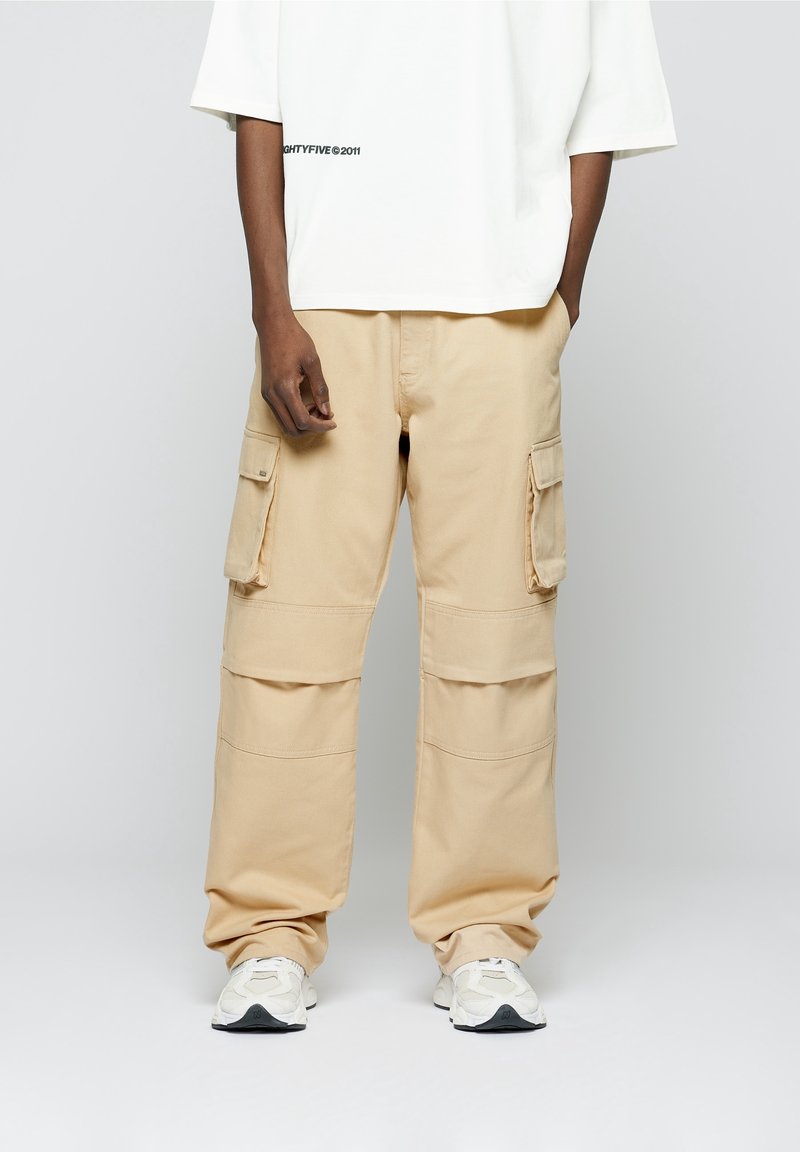 Pantalon cargo beige avec deux poches latérales, coupe ample, coutures renforcées et texture douce en coton, associé à des baskets blanches.