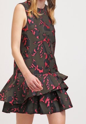 Femme portant une robe noire sans manches avec un motif floral abstrait rouge et une jupe à volants superposés, debout devant un fond uni.