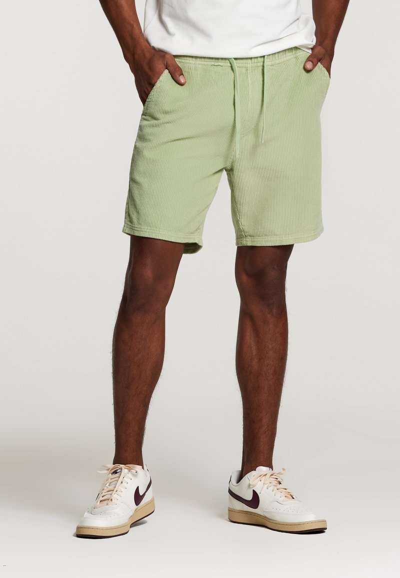 Shorts en velours côtelé vert clair avec une taille élastique, deux poches latérales et une coupe décontractée. Associés à des baskets blanches et un t-shirt.