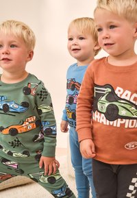 Trois enfants portent des tenues colorées : un ensemble vert avec des imprimés de voitures, un haut bleu avec un dessin de voiture, et une chemise orange avec l'inscription « CHAMPION DE COURSE ».