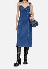 Kvinna iförd en blå denim ärmlös midiklänning, svarta knähöga klackstövlar, ett kedjehalsband och bär en denimaxelväska.