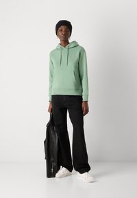 Ljusgrön huvtröja med framficka, tillsammans med svarta jeans och vita sneakers. Modellen håller en svart läderjacka.