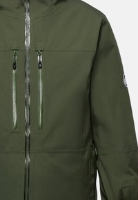 Giacca impermeabile verde con zip frontale completa, due tasche sul petto e un patch con il logo sulla manica sinistra. Ha una texture liscia.