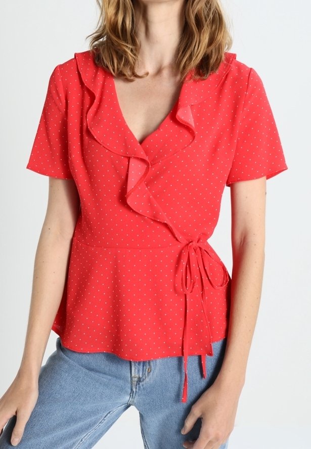 Femme portant un chemisier rouge à manches courtes avec petits pois blancs et col à volants, associé à un jean bleu clair.