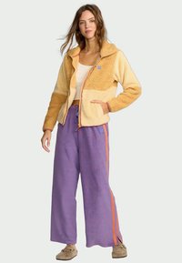 Casaco de felpa bege e creme, confortável, com fecho éclair, combinado com calças roxas largas que têm riscas laranja nas laterais. Textura suave em toda a peça.