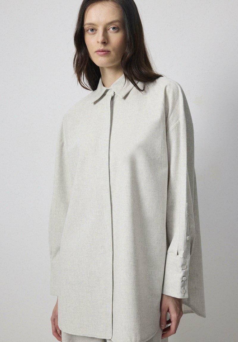 Chemise oversize gris clair à boutons, avec un col pointu, des manches longues avec des poignets à boutons, et un tissu à texture douce.