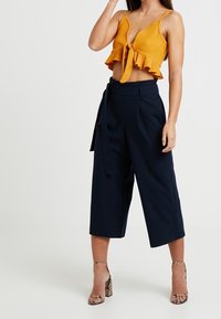Top de cultivo acanalado mostaza con dobladillo con volantes y detalle de nudo delantero, combinado con culottes de tiro alto color navy y sandalias de tacón con tiras.
