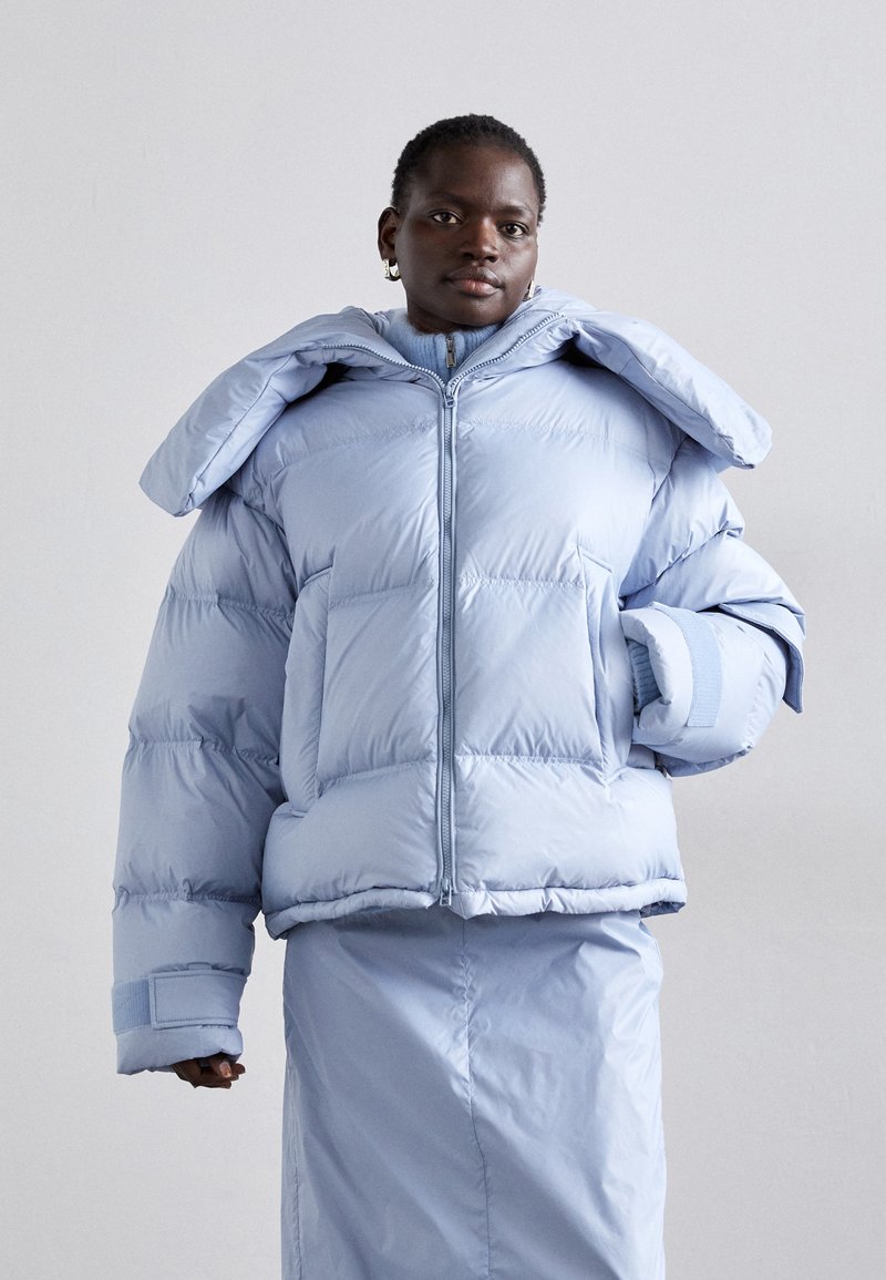 Lichtblauwe pufferjack met een oversized capuchon, lange mouwen en brede manchetten; heeft een gequiltde textuur en een frontale rits. Gecombineerd met een bijpassende rok.