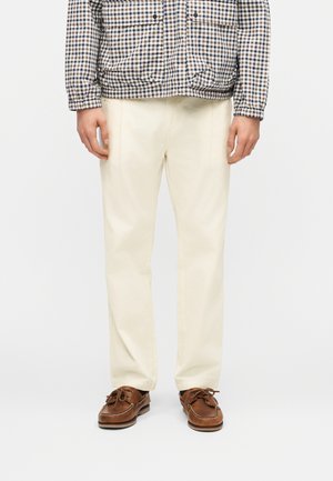 Moški, oblečen v kremaste ravne hlače, rjave usnjene čolnarice in karo jopič, stoji pred belim ozadjem.