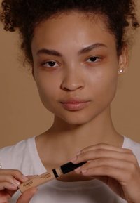 KIKO Concealer in einer beigen Tube mit schwarzem Deckel, von einer Person vor einem hellbraunen Hintergrund gehalten, die ein weißes Oberteil trägt. Glatte Hautstruktur sichtbar.