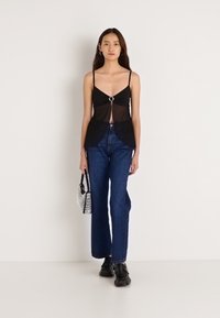 Melnas caurspīdīgas blūzes augšdaļa ar priekšējo mezglu un apaļiem metāla elementiem, valkāta kopā ar augsta vidukļa zilām džinsa biksēm un melniem apaviem, turot mazu sudraba somiņu.