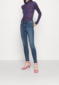 Jeans skinny azules con bolsillos delanteros, combinados con una blusa de mangas largas de patrón púrpura y sandalias de tacón alto rosas, mostrando una silueta esbelta.
