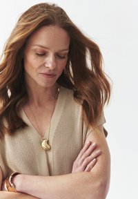 Suéter de punto beige con escote en V, llevado con un collar de pendiente dorado y una pulsera de metal. El collar presenta un diseño curvado en forma de riñón.