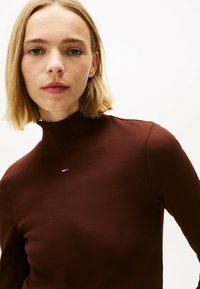Top marrone a collo alto a costine con un piccolo logo ricamato sul petto. Il design è aderente e presenta maniche lunghe.