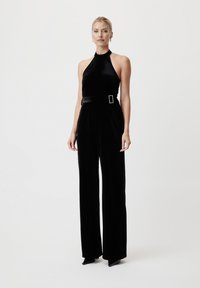 Svart fløyels jumpsuit med halterneck-design, vide ben og et svart belte med firkantet spenne. Glatt tekstur, formtilpasset silhuett.