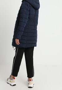 Marinblå pufferjacka med huva, med quiltat mönster, justerbar dragsko i midjan och elastiska ärmslut, bärs över svarta träningsbyxor med vita ränder.