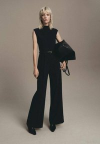 Mono negro con cuello alto, piernas anchas y cintura ceñida con cinturón. La modelo sostiene un bolso negro estructurado y lleva zapatos negros de punta.