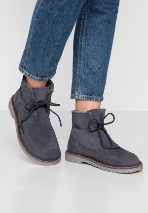 Personne portant des bottes à lacets en suede gris avec doublure en fourrure et semelles en liège, associées à un jean en denim bleu.