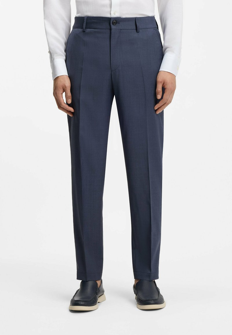 Homme portant un pantalon bleu marine sur mesure, une chemise blanche et des chaussures à enfiler bleu marine, debout devant un fond uni.