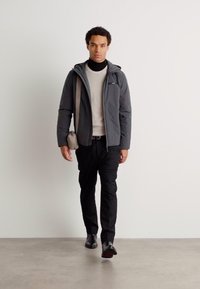 Giacca grigia con zip sopra un maglione chiaro, pantaloni neri e scarpe nere. Una piccola borsa a tracolla completa l'outfit. Sfondo neutro.