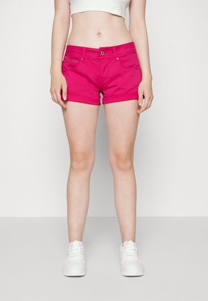 Mujer con pantalones cortos rosa brillante, top corto blanco y zapatillas blancas, posando frente a un fondo blanco liso.