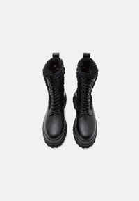 Een paar zwarte leren veterschoenen met dikke zolen en gekruld bont rondom de enkels, van bovenaf bekeken op een witte achtergrond.