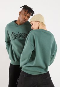 Felpa verde con la scritta "Visionary" in rilievo nera, caratterizzata da un colletto girocollo e una vestibilità ampia. Indossata da due modelli, uno dei quali con un berretto beige.