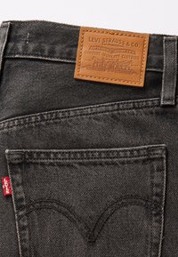 Les jeans en denim noir présentent une étiquette en cuir avec un logo en relief et une étiquette rouge sur la poche arrière. Des coutures détaillées mettent en valeur le design.