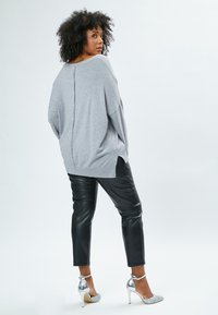 Maglione grigio oversize con dettaglio dei bottoni sul retro, abbinato a pantaloni in ecopelle neri e scarpe con tacco alto argento.