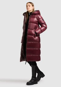 Femme portant un long manteau matelassé bordeaux brillant avec une capuche, des leggings noirs et des bottes noires à bout chunky arrivant au genou, se tenant devant un fond gris.