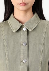 Oliivroheline teksashirt kraega, millel on ees hõbedased nööp-snapid ja tekstureeritud kangas vertikaalsete õmblustega.