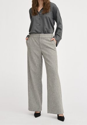 Woman wearing grey button-up shirt, wide-leg light grey trousers, black pointed-toe heels, standing with hands in pockets.