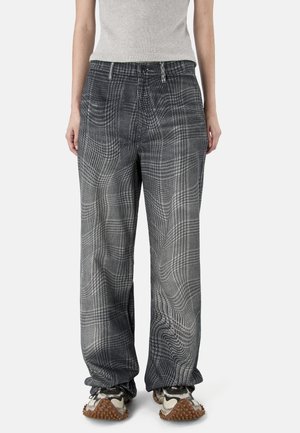 Osoba ubrana w luźne szare spodnie w kratę, jasnoszary top i masywne sneakersy z teksturowaną brązową podeszwą, stojąca na jednolitym tle.