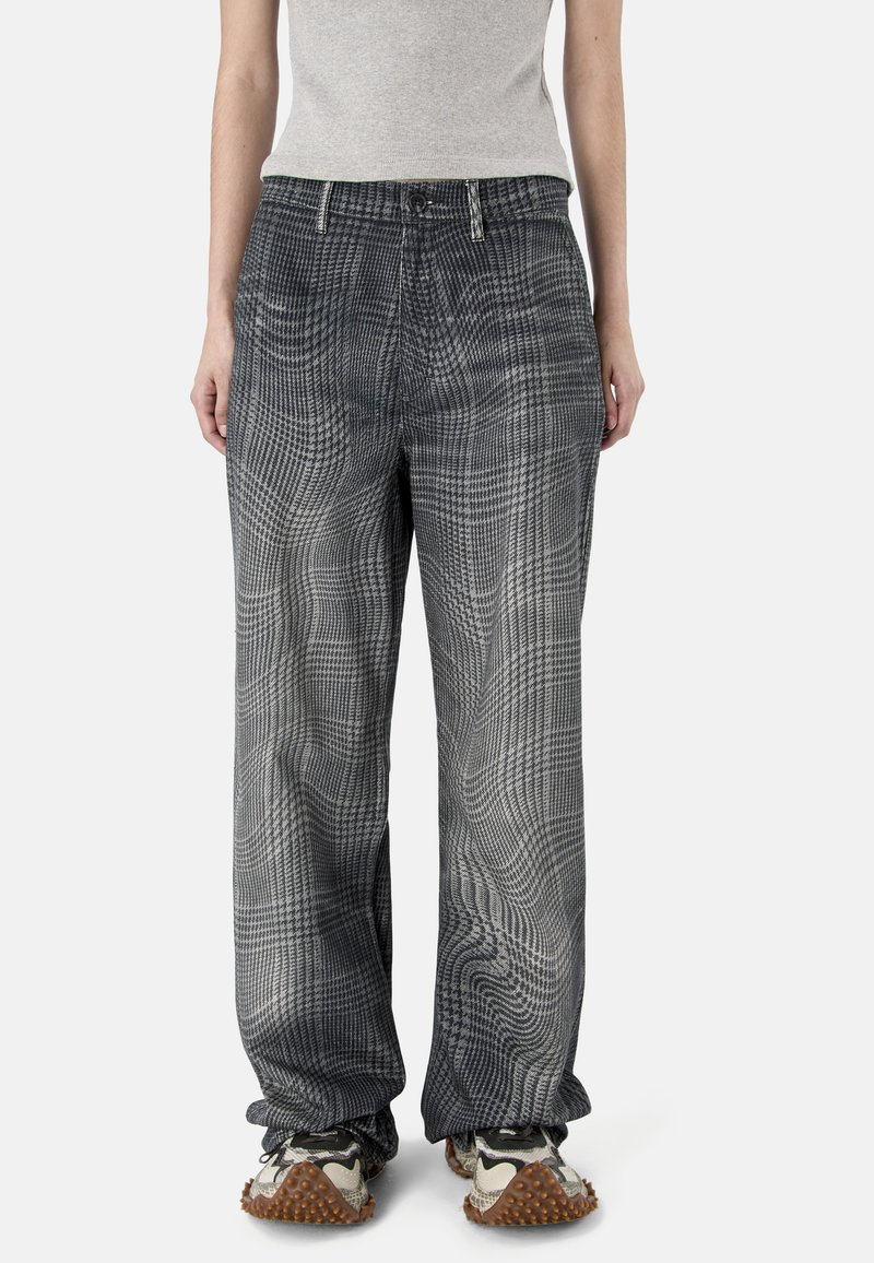 Persona vistiendo pantalones sueltos de cuadros grises, camiseta gris claro y zapatillas gruesas con suelas marrones texturizadas, de pie frente a un fondo liso.