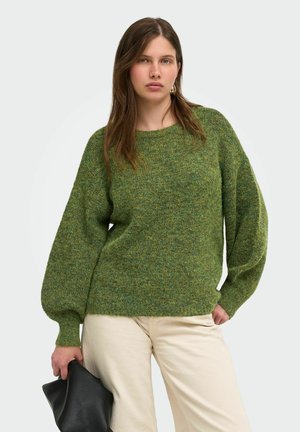 Femme portant un pull texturé vert et un pantalon beige clair tenant une pochette en cuir noire sur un fond uni.