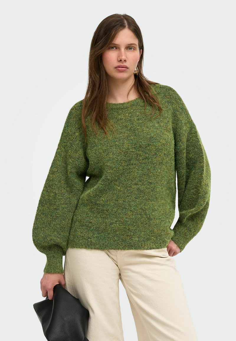 Femme portant un pull texturé vert et un pantalon beige clair tenant une pochette en cuir noire sur un fond uni.