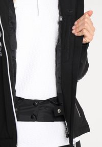 Czarna kurtka z zamkiem błyskawicznym z przodu, z fakturowaną białą warstwą wewnętrzną, bocznymi kieszeniami i zatrzaskami przy talii. Wykończona szarymi detalami.