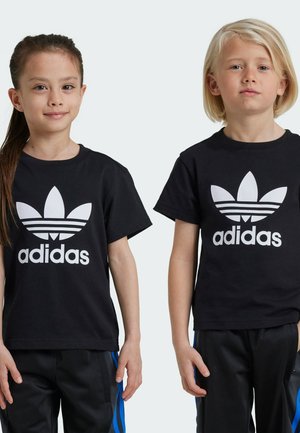 Des t-shirts en coton noirs présentent un grand logo Adidas blanc et un design en trèfle sur le devant, portés avec un pantalon noir à rayures bleues.