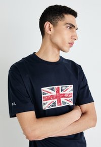 Navy zils kokvilnas T-krekls ar nolietotu Lielbritānijas karoga grafiku priekšā un logo uz kreisā piedurknes. Īsas piedurknes, apaļas kakla dizains.